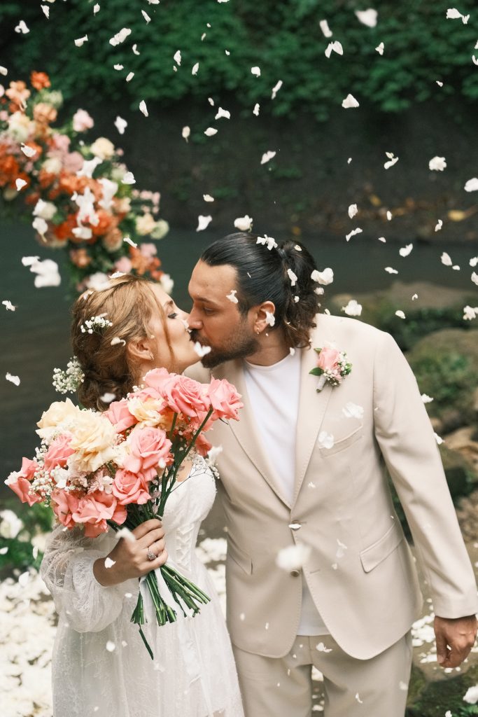 Flying white petals surrounding couple during kiss in front of a waterfall wedding florist bali setup